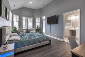 A bedroom with a bed, a TV, a desk, a chair, and a bathroom with a mirror and lights. - Modern Soundfront - Sleeps 12 (Manteo)