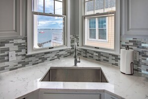 A kitchen with a large sink, two windows with views of the sea, and a tiled backsplash.