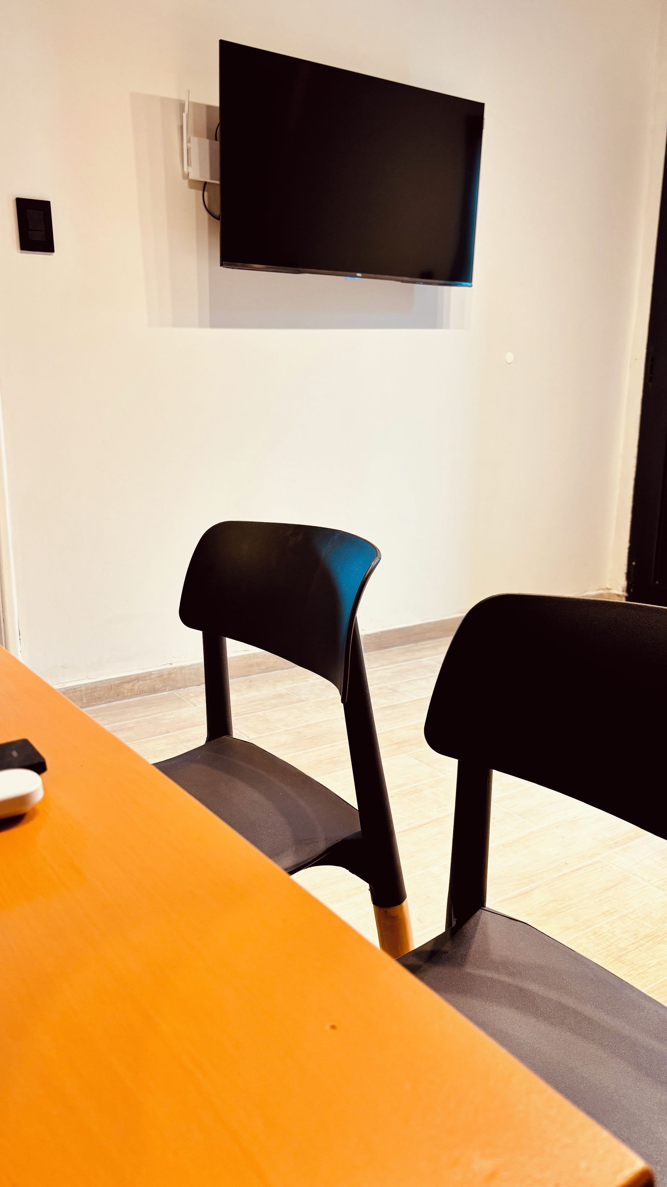 Una habitación con una televisión de pantalla plana montada en la pared, dos sillas negras y una mesa naranja.