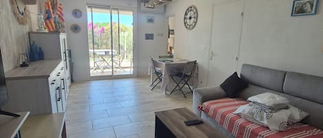 A spacious living area with a sofa, a coffee table, and a dining table. There is a sliding glass door leading to an outdoor area.