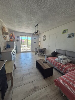 A spacious living area with a sofa, a coffee table, and a dining table. There is a sliding glass door leading to an outdoor area.