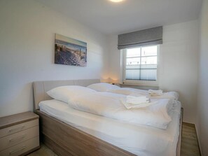 A neatly made bed with white linens and a folded towel on the side, a bedside table with drawers, a window with blinds, and a painting on the wall. - Apartment 14 (St. Peter-Ording)