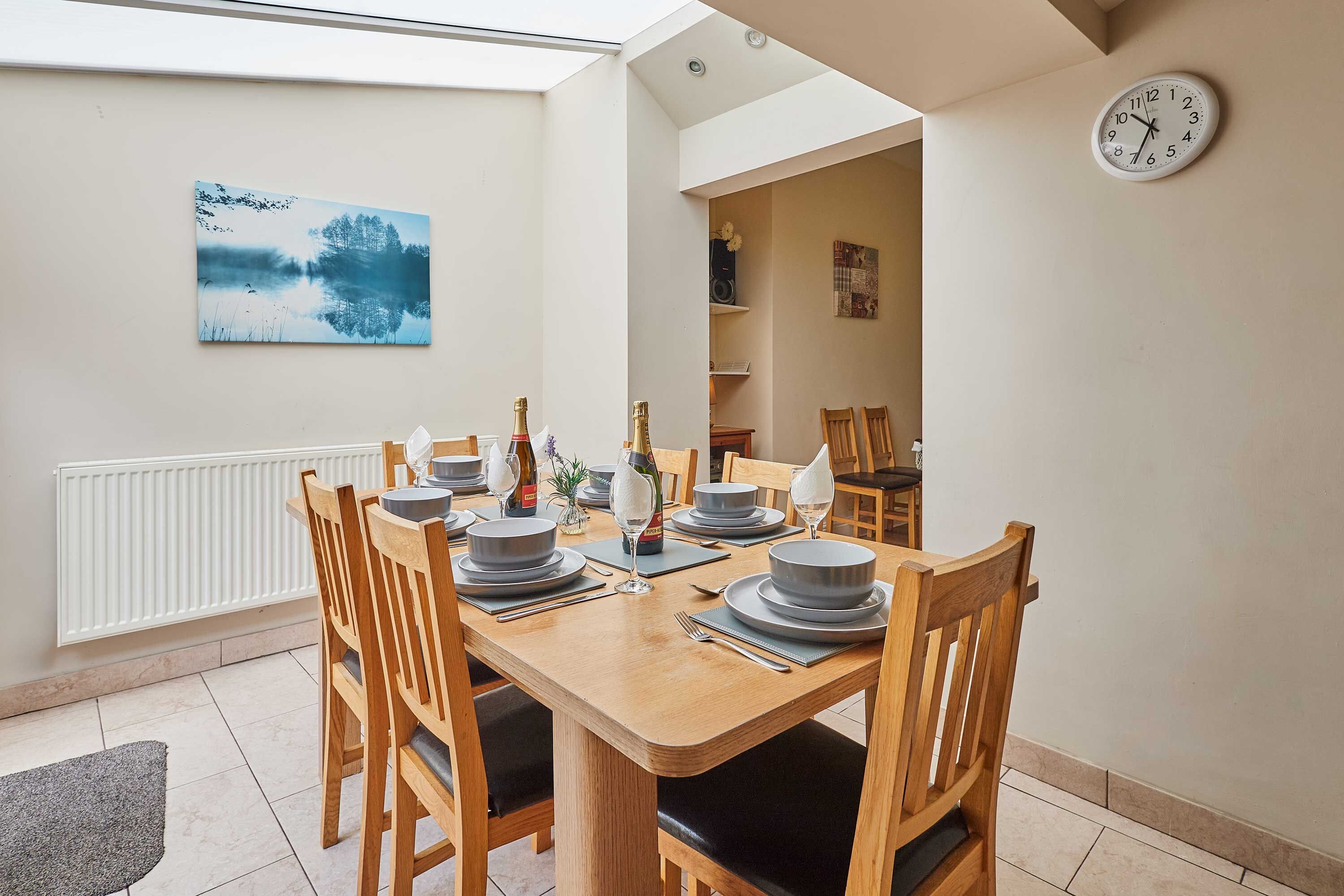 A dining area with a wooden table set for four, chairs, a clock on the wall, and a painting above the table.