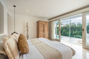 Ein Schlafzimmer mit einem großen Bett, einem Holzschrank und Blick auf eine tropische Landschaft durch große Fenster.