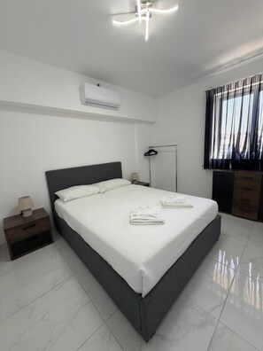 A modern hotel room with a large bed, a bedside table with a lamp, an air conditioning unit, a wardrobe, and a window with curtains. - Modern Apartment in Paceville Steps from Beach (San Ġiljan)