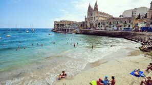 A beach with clear water, people swimming, and a historic building in the background. - Modern Apartment in Paceville Steps from Beach (San Ġiljan)