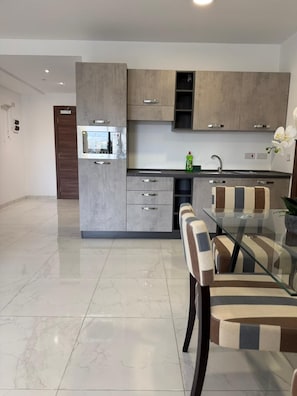 A modern kitchen with grey cabinetry, a built-in microwave, and a glass dining table with striped chairs. - Modern Apartment in Paceville Steps from Beach (San Ġiljan)