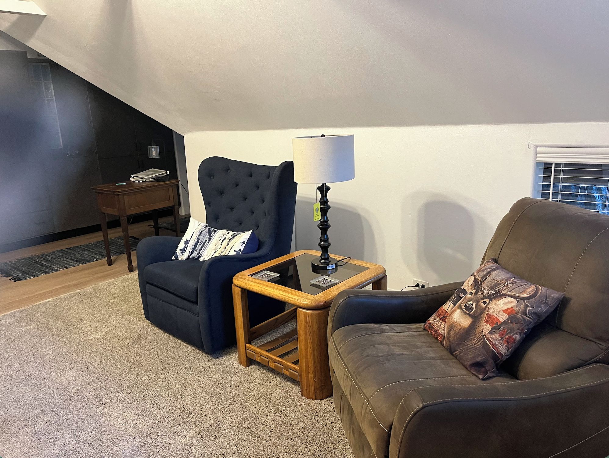 Un salon avec un fauteuil bleu foncé, un canapé marron, une petite table d’appoint en bois avec une lampe et un tapis.