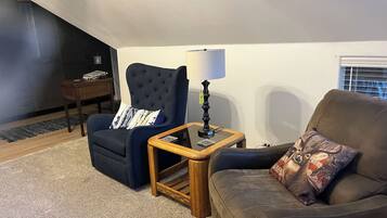 Un salon avec un fauteuil bleu foncé, un canapé marron, une petite table d’appoint en bois avec une lampe et un tapis.