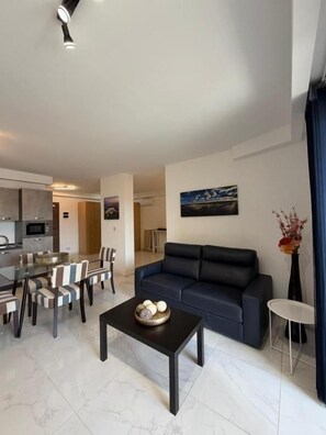 A modern living room with a dark sofa, a dining table with chairs, a kitchen area in the background, and a painting on the wall. - Modern Apartment in Paceville Steps from Beach (San Ġiljan)