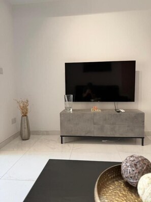 A modern living room with a flat-screen TV mounted on a grey cabinet, a glass vase with dried plants, and a black exercise mat. - Modern Apartment in Paceville Steps from Beach (San Ġiljan)