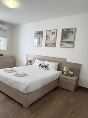 Cama bem arrumada com lençóis brancos e um travesseiro em formato de estrela-do-mar, mesa de cabeceira com abajur, e parede com três quadros emoldurados.