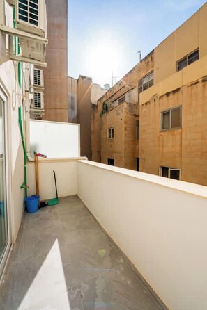 Ein schmaler Balkon mit Betonboden, einem weißen Geländer und Blick auf benachbarte Gebäude.