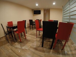 Un área de comedor con varias mesas y sillas, un televisor de pantalla plana montado en la pared y un separador de vidrio en un lateral.