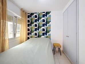 A bedroom with a bed, a window with curtains, a wall with a geometric pattern, a small stool, and a wardrobe. - DRASSANES Rosas / Roses – INTERimmo (Rosas / Roses)