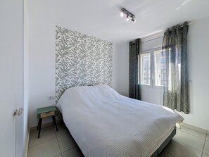 Ein Schlafzimmer mit einem Bett, einem kleinen Tisch, einer Wand mit Muster, einem Fenster mit Vorhängen und einer Deckenleuchte.