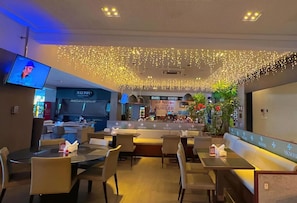 A restaurant with multiple tables and chairs, a bar area, and a television screen displaying a program. - Holandas Prime Studio Varanda Lateral (João Pessoa)