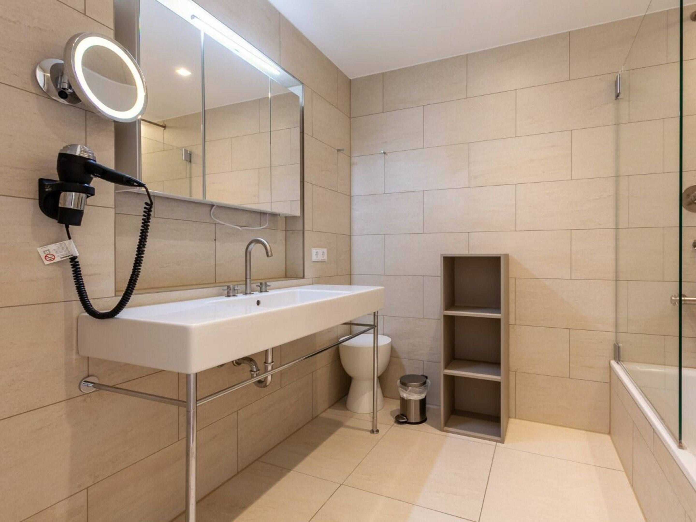 A modern bathroom with a white sink, a mirror with lights, a hairdryer, and a glass shower enclosure.