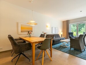 A modern living room with a dining area, featuring a wooden table, grey sofas, and a large window with a view of greenery. - Holiday Home With 1 Bedroom (St. Peter-Ording)