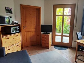 A compact room with a wooden door, a television on a wooden stand, a microwave on a shelf, a small table, and a chair. - Holiday Apartment With 1 Bedroom (Schwangau)