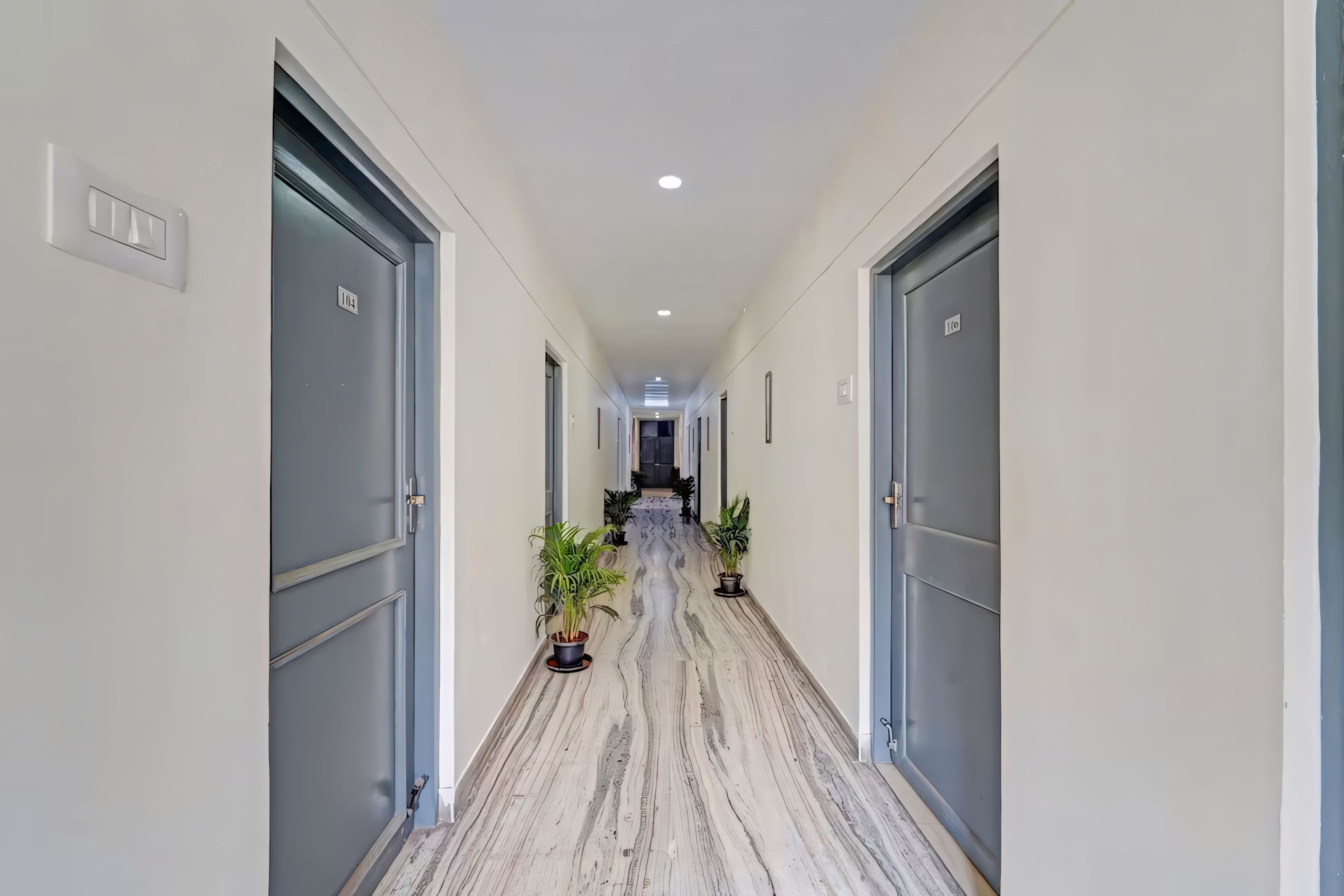 Corredor com portas numeradas 104 e 100, piso de mármore e plantas em vasos.