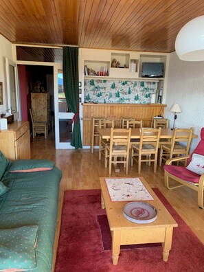 A cozy living room with a wooden ceiling, a green sofa, a red rug, a wooden coffee table with a plate, and a dining area with wooden chairs and a kitchen in the background. - 70-square-meter apartment with a view and ski-in/ski-out access right from the building (Huez)