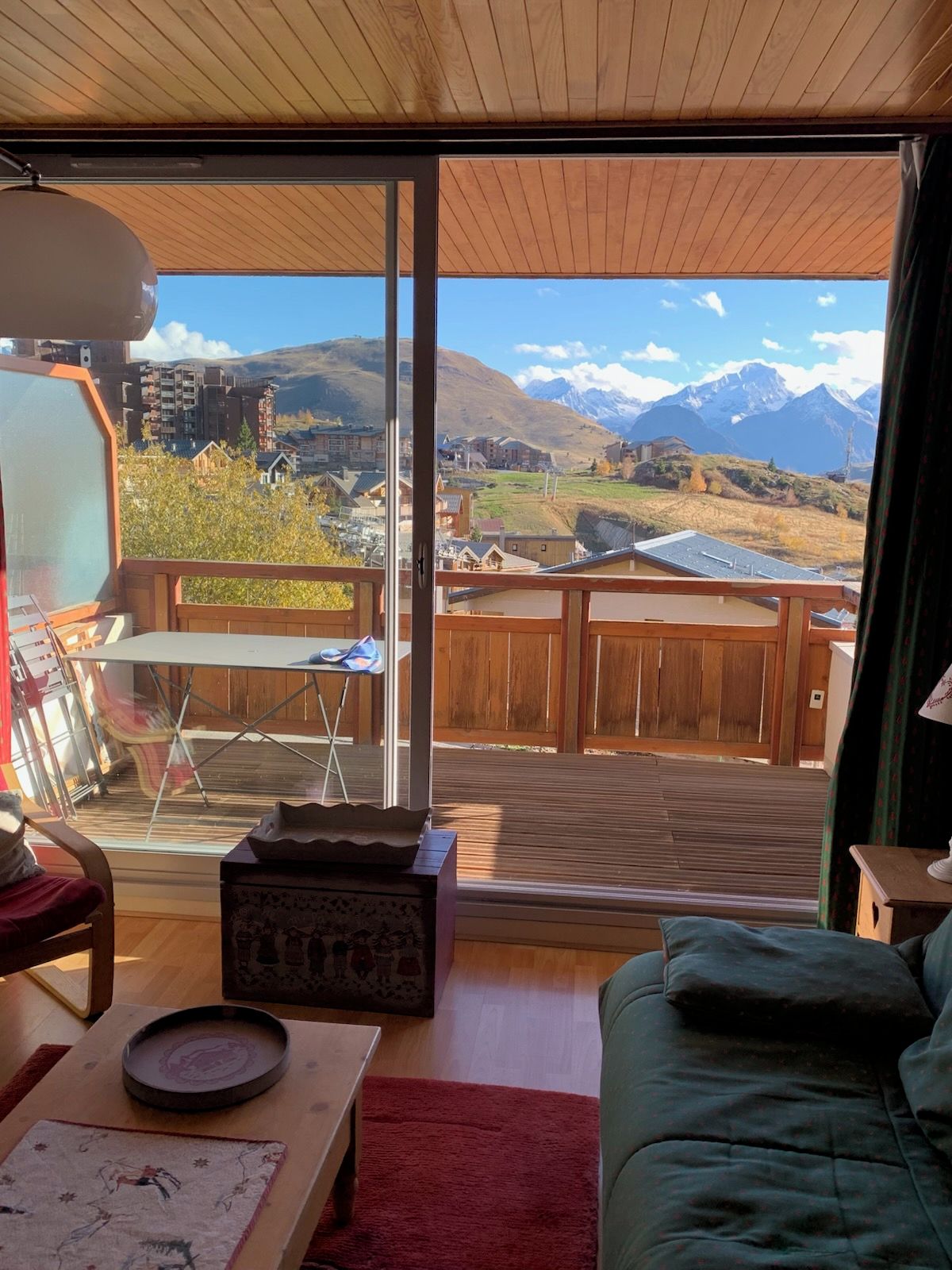 A room with a large window offering a mountain view, a wooden deck outside, and a cozy interior with a sofa and a table.