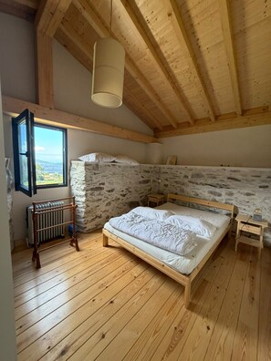 A bedroom with a stone wall, wooden ceiling beams, a bed, a radiator, and a window with a view. - Casa Lareira (Valdoviño)