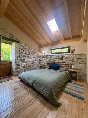 Une chambre avec un mur en pierre, des poutres en bois au plafond, un lit avec une literie verte, une table de chevet, une fenêtre et une petite plante.
