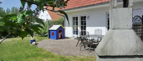Uma casa com telhado vermelho, um pequeno parquinho azul, área de churrasco e um pátio pavimentado.