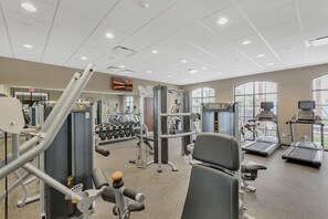 Un gimnasio con diversos equipos de ejercicio, incluyendo cintas de correr y máquinas de pesas, y grandes ventanales que proporcionan luz natural.