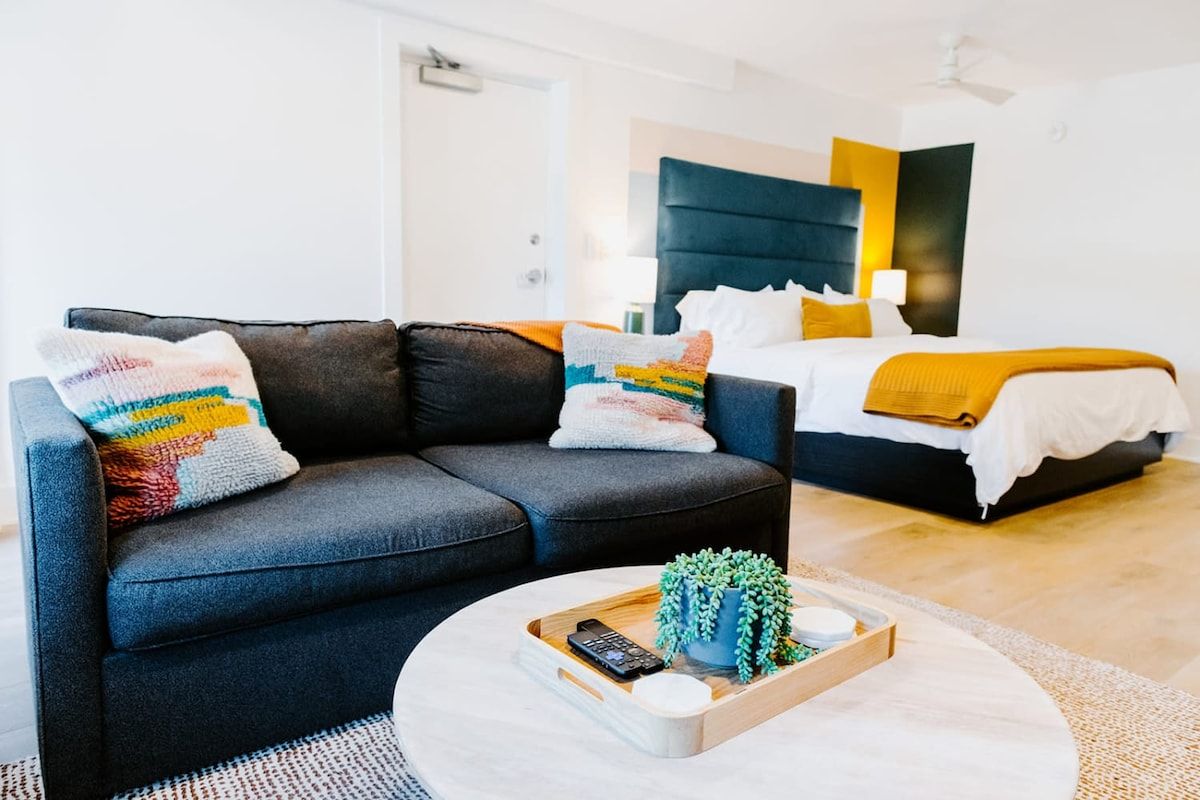 A modern hotel room with a dark-colored sofa, a round coffee table with a remote control and a plant, and a bed with a yellow blanket.