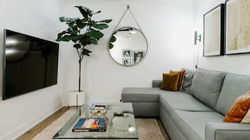 Una sala de estar moderna con un televisor de pantalla plana, un espejo redondo, una mesa de centro con libros, un sofá gris y una planta grande.