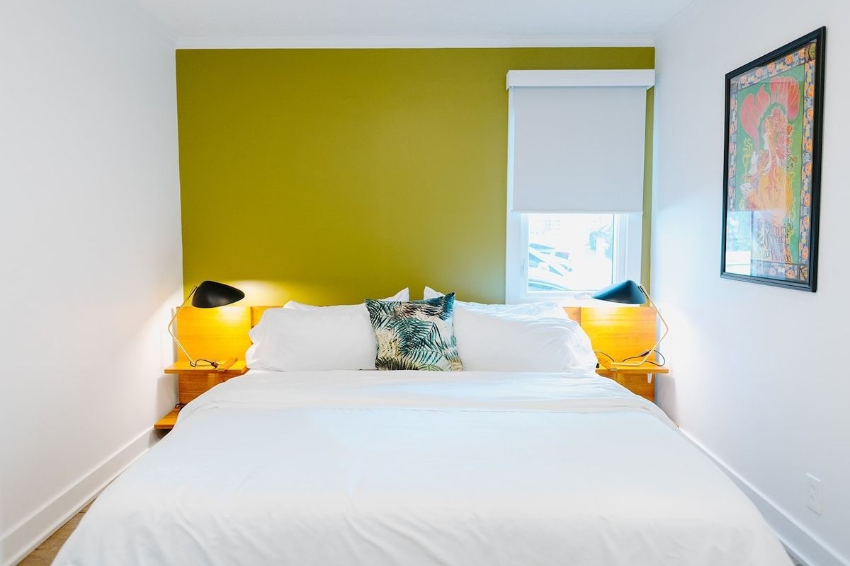 A bed with white bedding, a green accent wall, and a window with blinds.