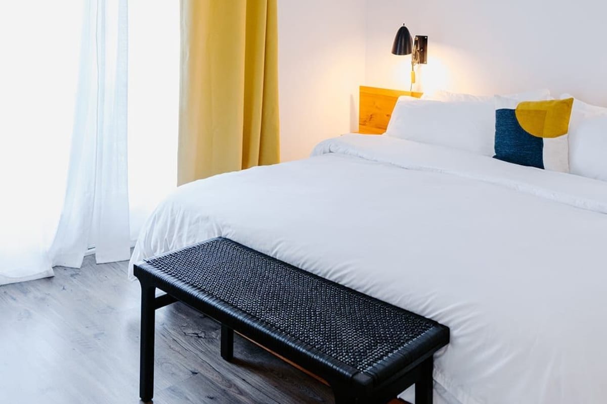 Una habitación de hotel con una cama, un banco, un ventilador de techo y una ventana con cortinas.
