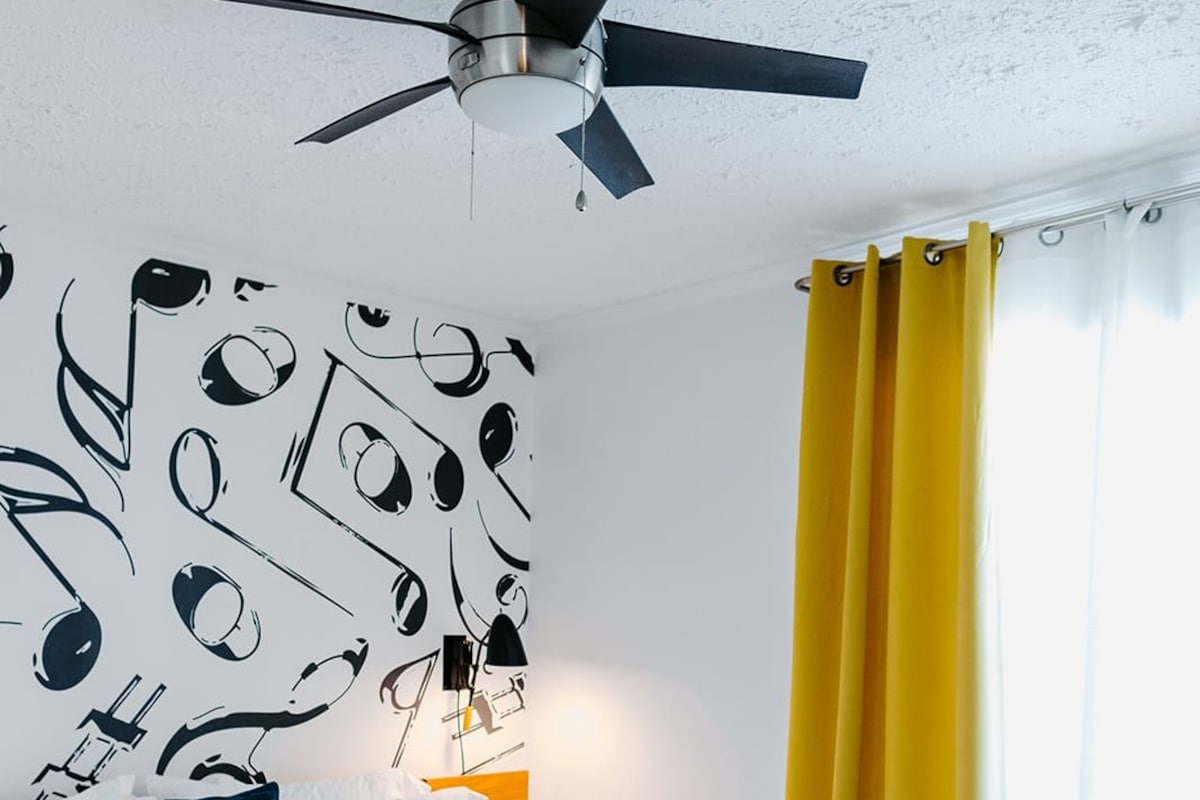 Un dormitorio con cama, ventilador de techo, pared con arte abstracto, mesita de noche con lámpara y ventana con cortinas.
