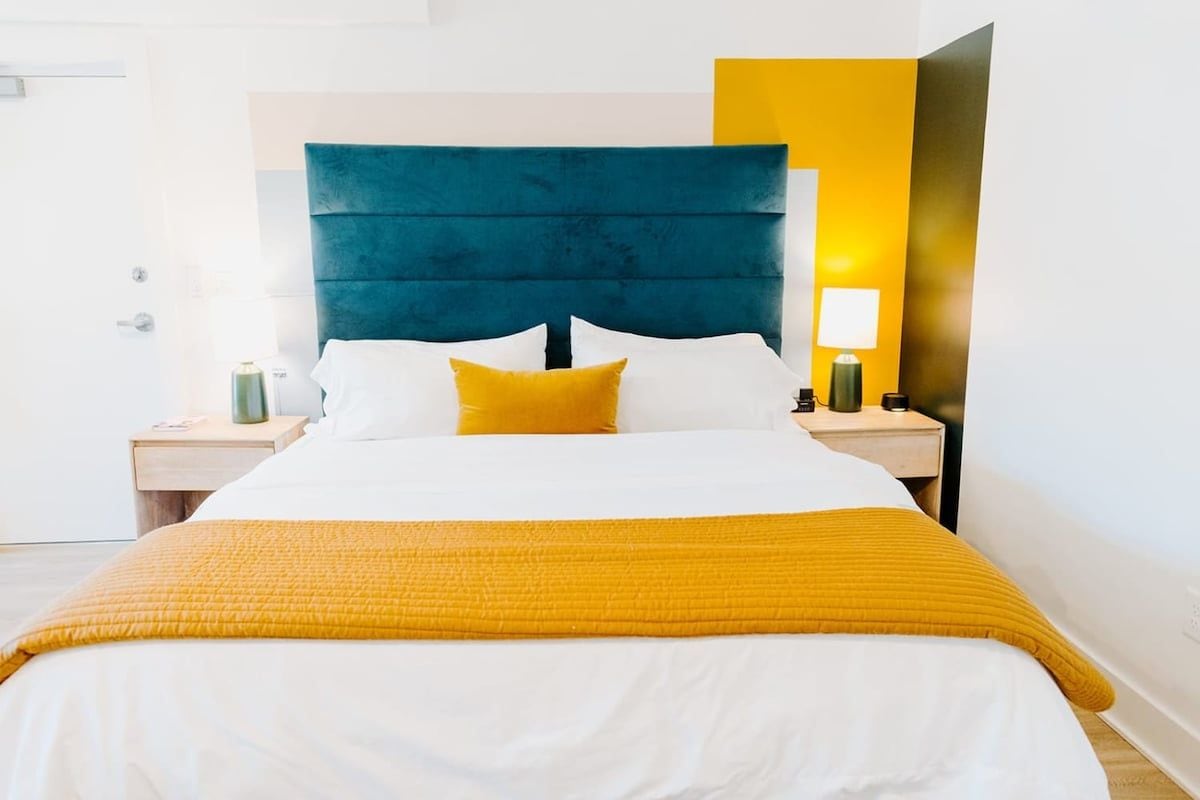 Una habitación de hotel con una cama grande, cabecero, mesita de noche, lámpara y aplique de pared.
