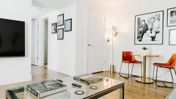 Una sala moderna con una mesa de centro de vidrio, un televisor de pantalla plana y una zona de comedor con dos sillas naranjas y una mesa redonda pequeña.