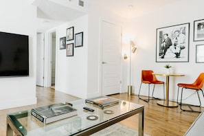A modern living room with a glass coffee table, a flat-screen TV, and a dining area with two orange chairs and a small round table.