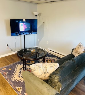 Una sala de estar con un televisor de pantalla plana montado en la pared, una mesa de centro de vidrio, un sofá oscuro con cojines estampados y una alfombra con motivos.