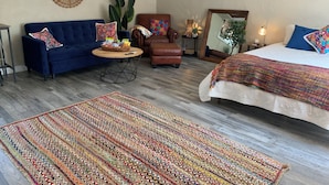 Een kamer met een blauwe bank, een bruine fauteuil, een bed met een kleurrijke deken, een houten salontafel en een kamerbreed tapijt met een patroon.