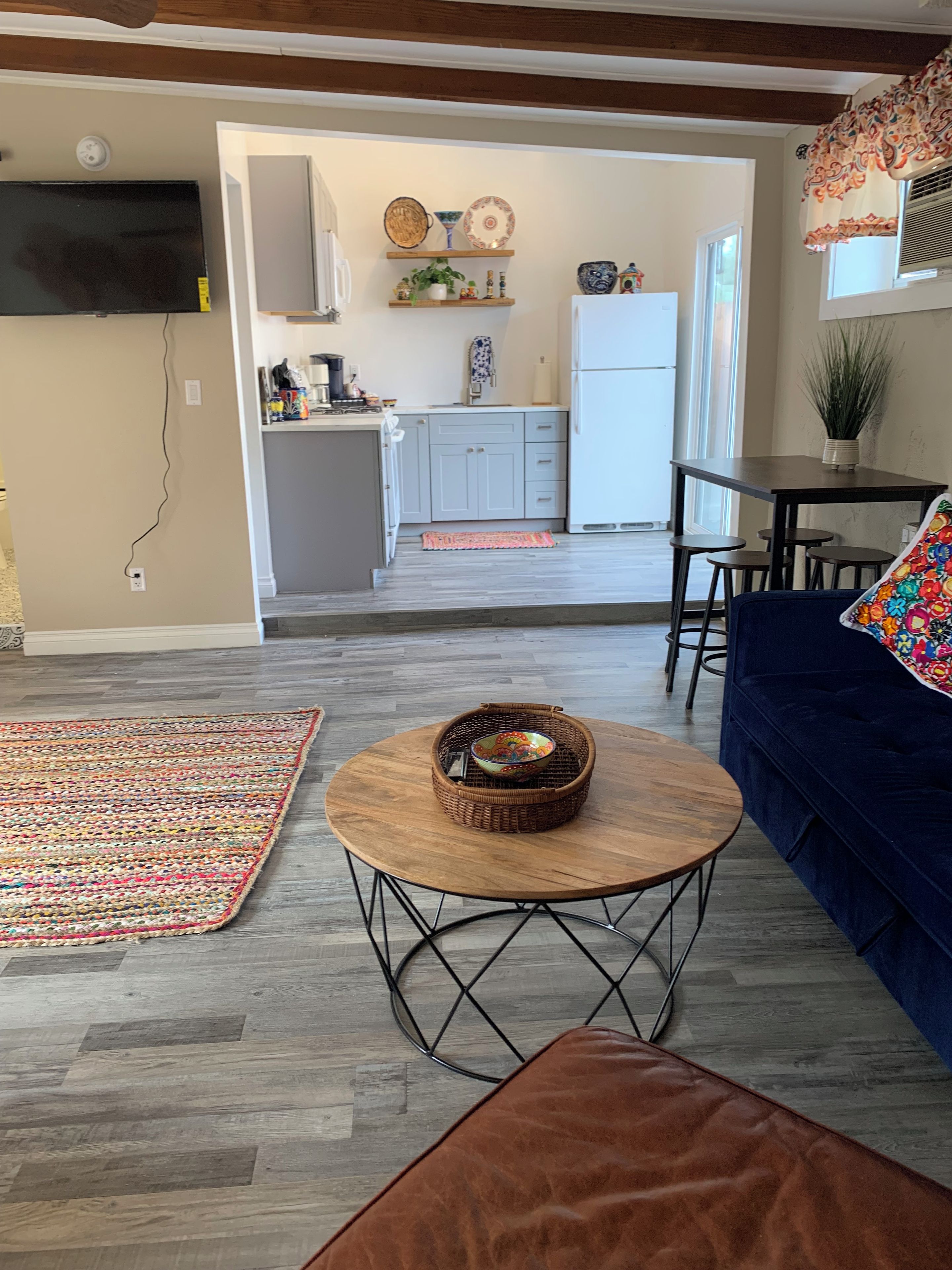 A living room with a sofa, a round coffee table with a woven basket, a patterned rug, and a kitchen area with cabinets and a refrigerator.