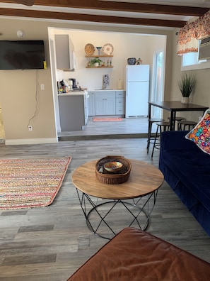 Un salon comprenant un canapé, une table basse ronde avec un panier en osier, un tapis à motifs et un coin cuisine avec des placards et un réfrigérateur.