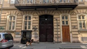 En bygning i klassisk arkitekturstil, med en hovedindgang prydet med en mørk trædør, en balkon med dekorativt metalgelænder og en lille hund foran.