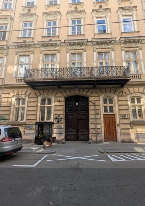 Ein Gebäude im klassischen Stil, mit einem zentralen Eingang, einer dunklen Holztür, einem Balkon mit aufwendiger Metallverzierung und einem kleinen Hund im Vordergrund.