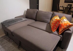 A grey sofa with a throw blanket and patterned pillows in a room with a door and a dining area in the background. - Cozy 2 bedroom house in Trincity (Trincity)