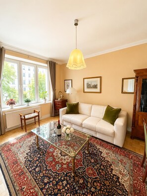 Ein Wohnzimmer mit einer Couch, einem Glastisch, einem gemusterten Teppich, einer Hängelampe und Blick auf Gebäude durch große Fenster.