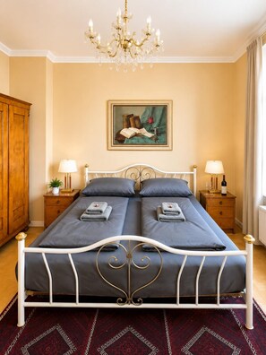 Ein Doppelbett mit weißem Metallgestell, zwei Nachttische mit Lampen, eine Chandelüster, ein Bild an der Wand und ein Holzschrank.