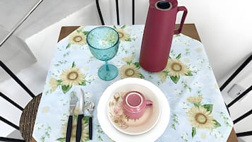 Uma mesa posta com toalha florida, caneca rosa, copo, garrafa térmica vermelha e talheres.