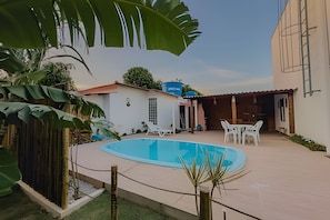 A swimming pool with a patio area, surrounded by a white building with a red roof, and a blue water tank in the background. - Apartamento 3 com 2 suítes 450m da praia (Santa Cruz Cabrália)
